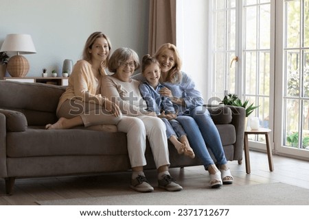 Happy girls and women of four different generations resting close on home sofa, smiling, laughing, looking at camera. Girl, mother, grandma, great grandmother full length portrait