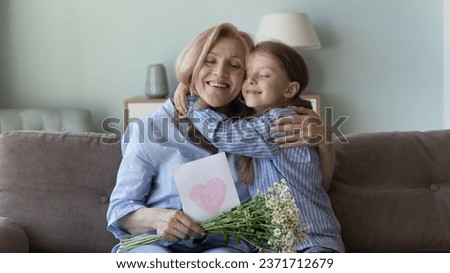 Cheerful sweet grandkid girl hugging happy grateful grandmother on sofa with love, congratulating on birthday, 8 march. Grandma holding handmade drawn card with heart, flowers, smiling