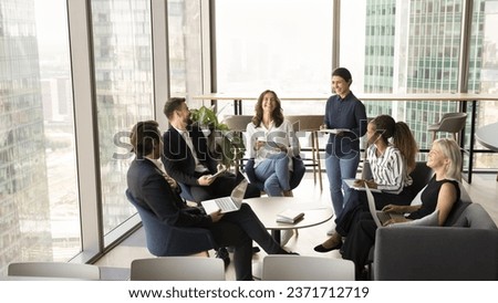 Happy diverse group of business colleagues laughing on meeting in office lobby, modern co-working space, sitting on couch, talking, discussing cooperation, professional tasks