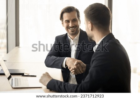 Happy young businessmen in formal jackets shaking hands at workplace table after meeting, discussing deal, hiring, business cooperation. Colleagues giving greeting handshakes Royalty-Free Stock Photo #2371712923
