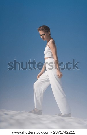 Caucasian adult woman tourist walking through snow with light fog on sunny weather with blue sky. Stylish blond female looking down, dressed in white cropped top, white pants and sandals. Full length