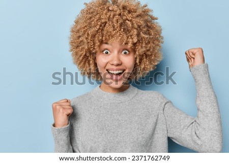 Happy attractive curly haired woman clenches fists with triumph and exclaims loudly feels like winner dressed in casual grey jumper isolated over blue wall. Cheerful female model makes winner gesture