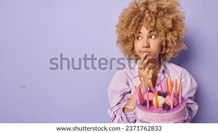 Horizontal shot of beautiful curly haired woman keeps hand on chin concentrated aside poses with tasty cake and burning candles celebrates special occasion isolated over purple background copy space Royalty-Free Stock Photo #2371762833
