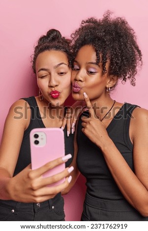 Vertical shot of two lovely women keep lips folded take selfie via smartphone apply makeup wear brisht makeup dressed in black t shirts isolated over pink background spend free time together