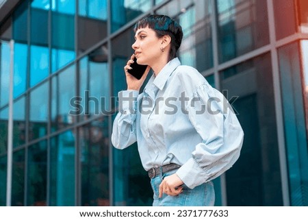 a woman in a blue shirt uses a mobile phone and a business center in the background Smiling lady professional holding cellphone standing at city street building looking away working outside office