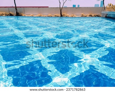 Blue water of swimming pool with square ceramics inside and trees around the pool. 