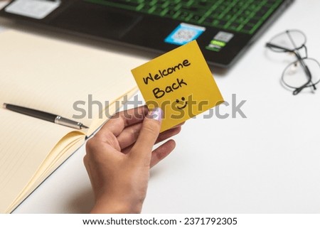 Human hand holding adhesive note with Welcome Back text on white background, laptop, diary, pen and spectacles in background