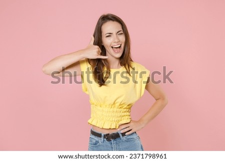 Blinking funny young brunette woman 20s wearing yellow casual t-shirt posing doing phone gesture like says call me back looking camera isolated on pastel pink color wall background studio portrait
