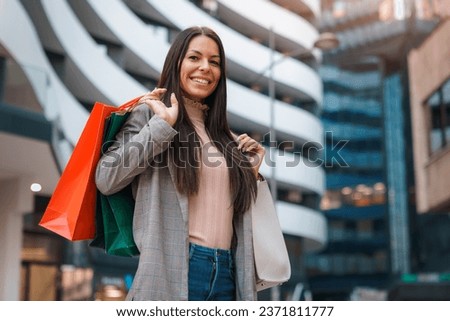 Beautiful young woman looking at camera wearing fashionable clothes holding shopping bags and purse while shopping in modern architectural ambient.