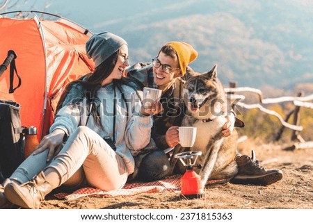 Photo of a happy young couple and their dog camping in the mountains on a beautiful autumn day and drinking a cup of tea outdoors.