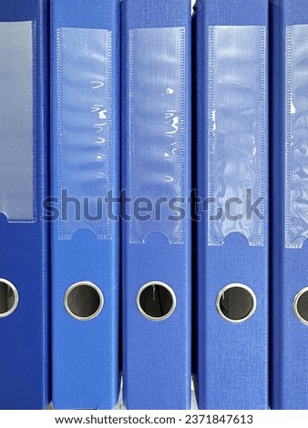 Blue A4 format binders for storing papers, standing on a shelf. In the lower part of the folder there is a round hole with a metal outline, on top there is a transparent pocket for signing the folder.