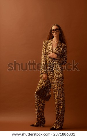 Fashionable confident woman wearing trendy leopard print satin suit blazer, trousers, sunglasses, holding small leather bag, posing in studio, on brown background. Fill-length portrait 