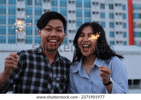 happy asian couple holding sparkler looking at camera with smiling face, celebrate new years eve, on urban building background, wear casual shirt in outdoor