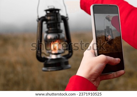 Woman photographs a kerosene lamp in her hand on a smartphone while on the field in the morning fog. A close-up photo from the first person.