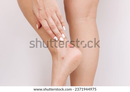 Callus on the heel from tight shoes. Cropped shot of a young woman touching her ankle with a sore with her hand on gray background. Corns, blisters on the foot Royalty-Free Stock Photo #2371944975