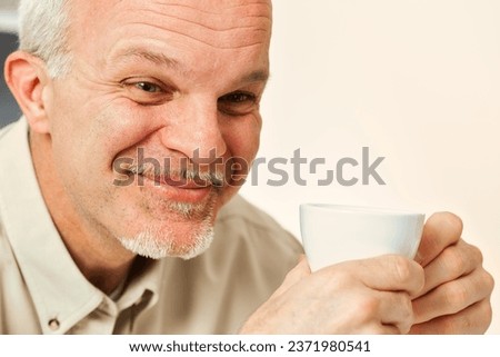 Amidst his thoughts, an aged man tries not to laugh. Holding a drink, likely coffee or tea, he's amused by the notion of something naive or laughable. His seasoned life offers such perspective Royalty-Free Stock Photo #2371980541