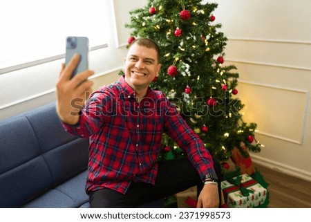 Attractive happy mexican man taking a selfie with her phone next to the christmas tree celebrating the holidays for social media