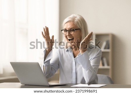 Cheerful blonde elderly business woman celebrating professional success, working at laptop, laughing, shouting for joy at home office workplace, getting good news from video call Royalty-Free Stock Photo #2371998691