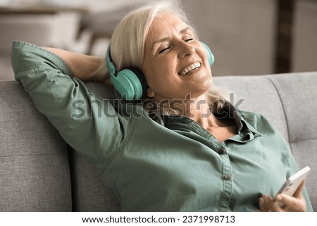 Cheerful inspired blonde elderly woman singing song at home, listening to music from big wireless headphones and online media service on cellphone, resting on sofa with closed eyes and toothy smile Royalty-Free Stock Photo #2371998713