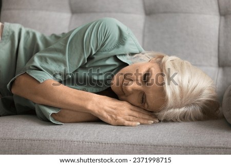 Sleepy napping blonde senior woman lying on side on comfortable couch, sleeping at home on break, enjoying leisure, calmness, caring for health, wellbeing Royalty-Free Stock Photo #2371998715
