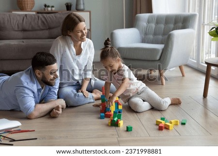 Happy attractive parents and little daughter kid stacking colorful toy blocks on heating floor together, building tower, relaxing at cozy home, ejoying family playtime, leisure Royalty-Free Stock Photo #2371998881
