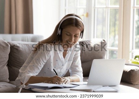 Happy pretty millennial woman using wireless headphones, laptop computer, watching learning webinar, lesson, talking on video call to business client, giving help to customer, writing notes