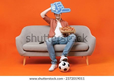 Young happy man football fan in shirt support team with soccer ball sit sofa at home watch tv live stream hold pizza hiding with foam glove finger isolated on orange background People sport concept.