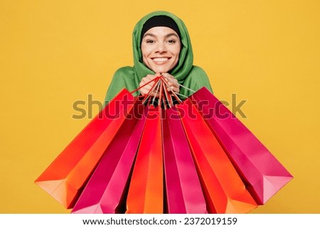 Young satisfied happy arab asian muslim woman wears green hijab abaya clothes hold shopping package bag look camera isolated on plain yellow background. Black Friday sale buy day, uae islam concept