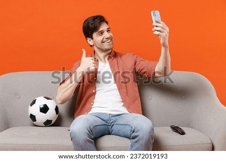 Young man football fan in shirt support team with soccer ball sit on sofa home watch tv live stream do selfie shot on mobile phone show thumb up isolated on orange background. People sport concept.