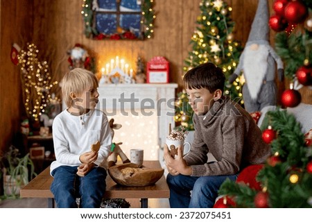 Beautiful blond child and his old brother, young school boys, playing in a decorated home with knitted toys at Christmas, eating cookies and drinking milk