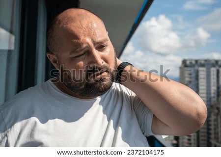 Bearded man in hot summer sun on balcony. Bald, unsmiling guy closed eyes in displeasure discontent from discomfort, holding on to sore neck, kneading ligaments, suffering from protrusion, pain, ache Royalty-Free Stock Photo #2372104175