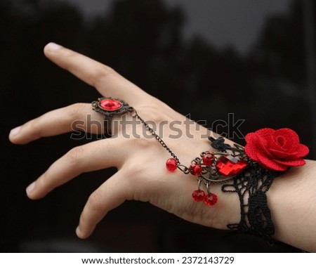 A feminine hand reaching out towards something. The hand is wearing black laced jewelry with red roses on it. Hand trying to grab something. Hand against dark background. Black background. Goth girl.