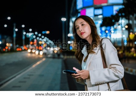 Close up of young Asian woman using mobile app device on smartphone to arrange taxi ride in city street. I Illuminated busy city traffic scene during rush hour with traffic congestion at night.