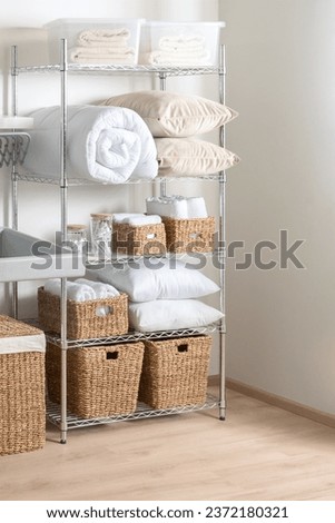 Interior of Bright Modern Laundry Room with White Duvet, Towels, Cozy Pillows, and Wicker Baskets on Adjustable Metal Storage Rack with Shelves, Chrome Wire Shelving Unit, Over Wooden Flooring. Royalty-Free Stock Photo #2372180321