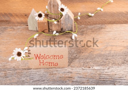Welcome home banner or note with white daisy flowers.  Tiny houses in background.
