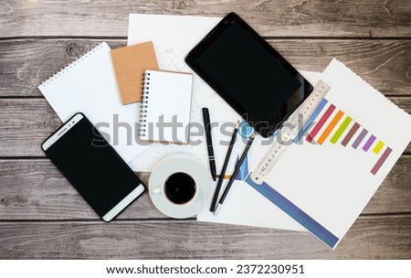 On a wooden background, an economist’s desk, a tablet, a notepad, pens, pencils, financial graphs, drawings, sheets of paper, a cup of coffee.  Office work concept, coffee break, flat lay top view.