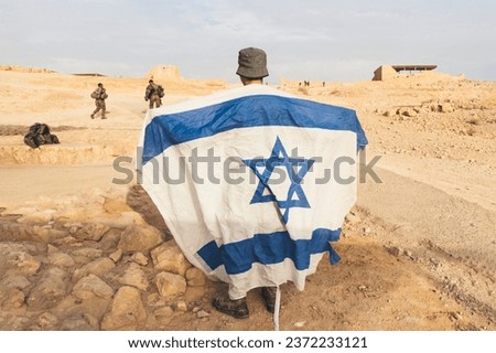 Back view of Israeli soldier with white and blue flag of Israel against the background of the desert and rocks.. Military men protecting promissed land. National flag of Israel. War conflict Royalty-Free Stock Photo #2372233121