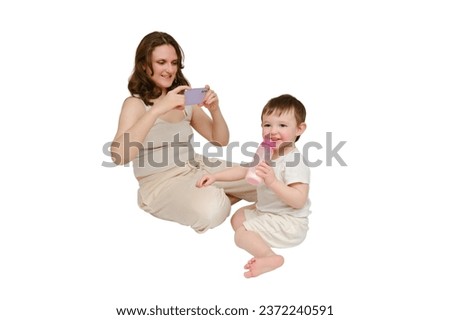 Woman mother takes a picture of a baby with a phone on a studio, isolated on white background. Mom blogger shoots a video from a child for a blog. Kid about two years old (one year nine months)