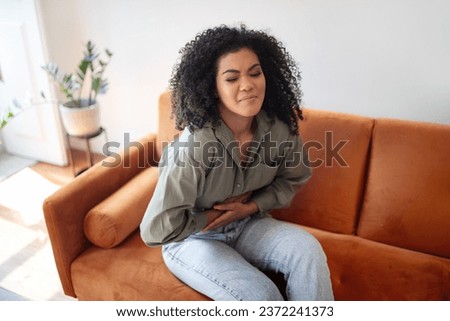 Gastritis Disease. Black woman suffering from stomachache, touching her stomach while having painful spasm in belly sitting on couch at home. Abdominal pain symptom, health problems concept Royalty-Free Stock Photo #2372241373