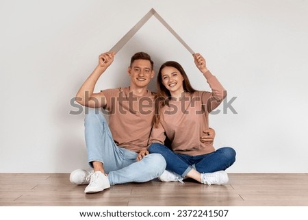 Real Estate Concept. Young Happy Couple Sitting Under Symbolic Cardboard Roof Near White Wall, Smiling Millennial Spouses Dreaming Of New Home, Planning To Buy Property, Copy Space Royalty-Free Stock Photo #2372241507
