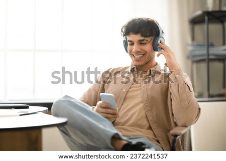 Glad Young Arab Guy Having Fun With Smartphone, Listens Online Music In Wireless Earphones, Sitting Near Desk During Work Day At Home Interior. Modern Gadgets Lifestyle Concept Royalty-Free Stock Photo #2372241537