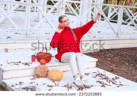 Beautiful woman is having a picnic in a beautiful place in autumn nature. She is holding and doing selfie with orange pumpkin. There are basket, book and coffee. Halloween concept. 