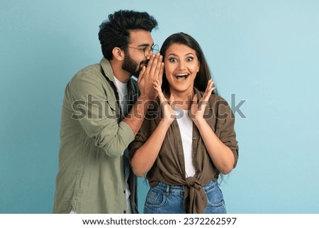 Handsome young middle eastern guy sharing secret or whispering gossips into his girlfriend's ear, excited pretty brunette indian woman gesturing, isolated on blue studio background, copy space Royalty-Free Stock Photo #2372262597