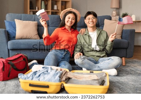 Cheerful Korean Couple Holding USA Flag and Packing Suitcase at Home, Smiling at Camera Showing Their Passports And Tickets. Young Spouses Preparing for Travel or Possibly Relocating Royalty-Free Stock Photo #2372262753