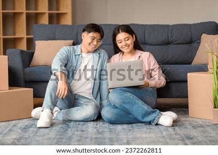 Asian Couple Using Laptop Websurfing And Searching New Rental Apartment, Using Online Service For Moving, Sitting Amid Packed Carton Boxes And Packages In Their Living Room At Home Royalty-Free Stock Photo #2372262811