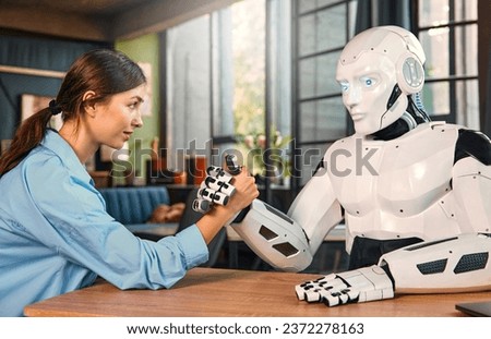 Young woman and humanoid robot doing arm wrestling while sitting at a table in a modern office. Collaboration between humans and artificial intelligence.