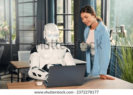 Young woman with a cup of coffee and a humanoid robot working while sitting at a laptop in a modern office. Collaboration between humans and artificial intelligence. Royalty-Free Stock Photo #2372278177