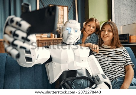 Mom with her daughter and a robot sitting on the sofa in the living room taking a selfie on a smartphone. Living together between humans and artificial intelligence. Royalty-Free Stock Photo #2372278443