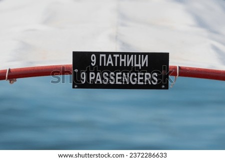 Nine passengers, only.  White letters,  on black board rectangle shape. Part of the boat. Information. White awning. Word and number on english language. 9 Patnici. On Macedonian language. Ohrid Lake.
