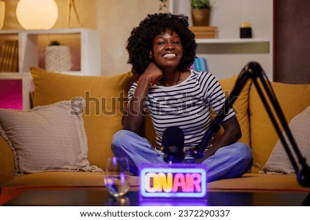 Young african american woman smiling while recording an audio broadcast in a modern cozy studio. Cheerful black female content creator hosting an internet podcast. Copy space. Looking into the camera.